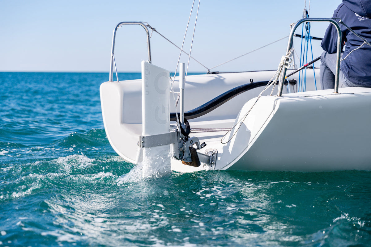 Close-up of a white sailboat's stern and outboard motor in turquoise ocean water under a clear blue sky, with part of a person visible on deck.