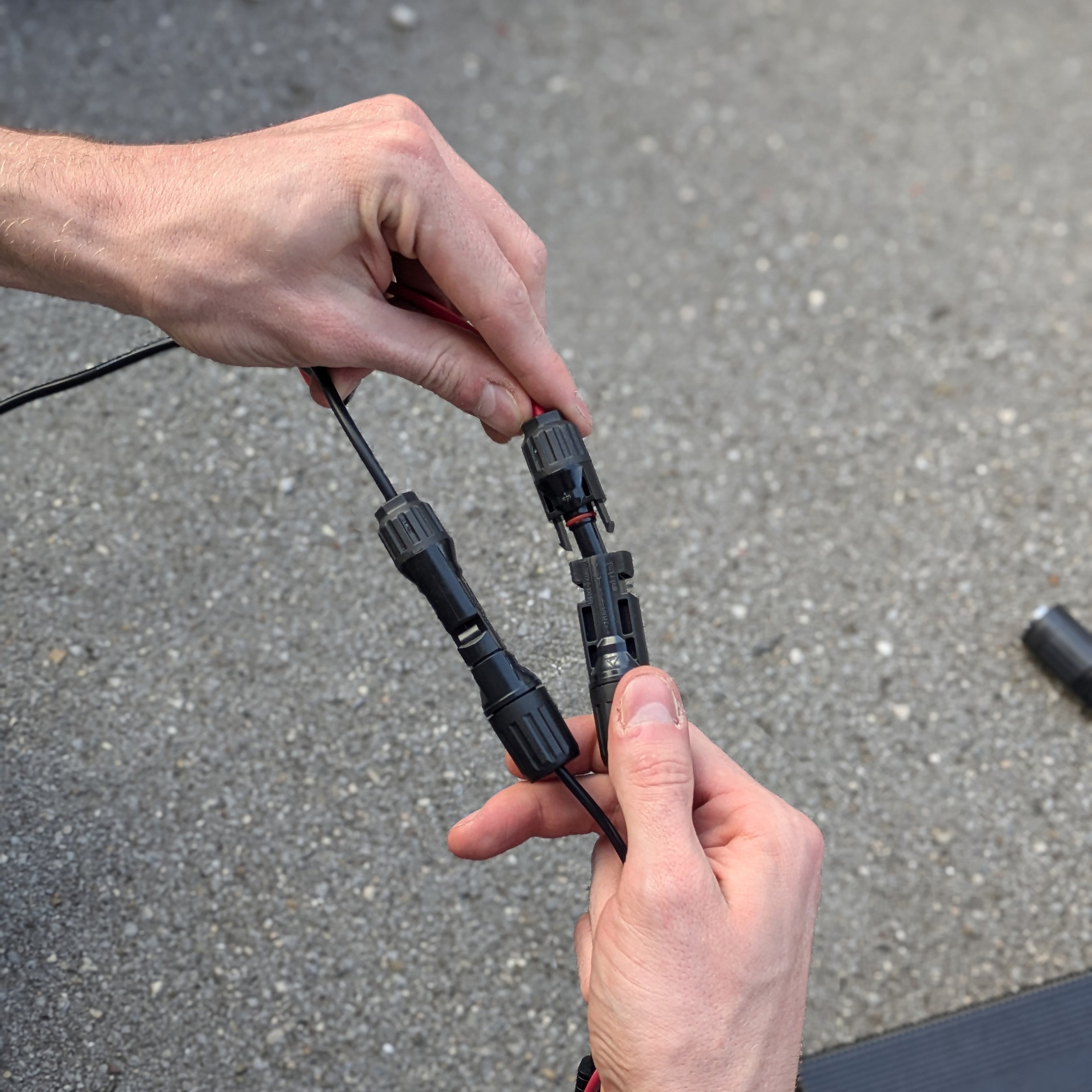 Two hands holding black electrical connectors against a gray pavement background
